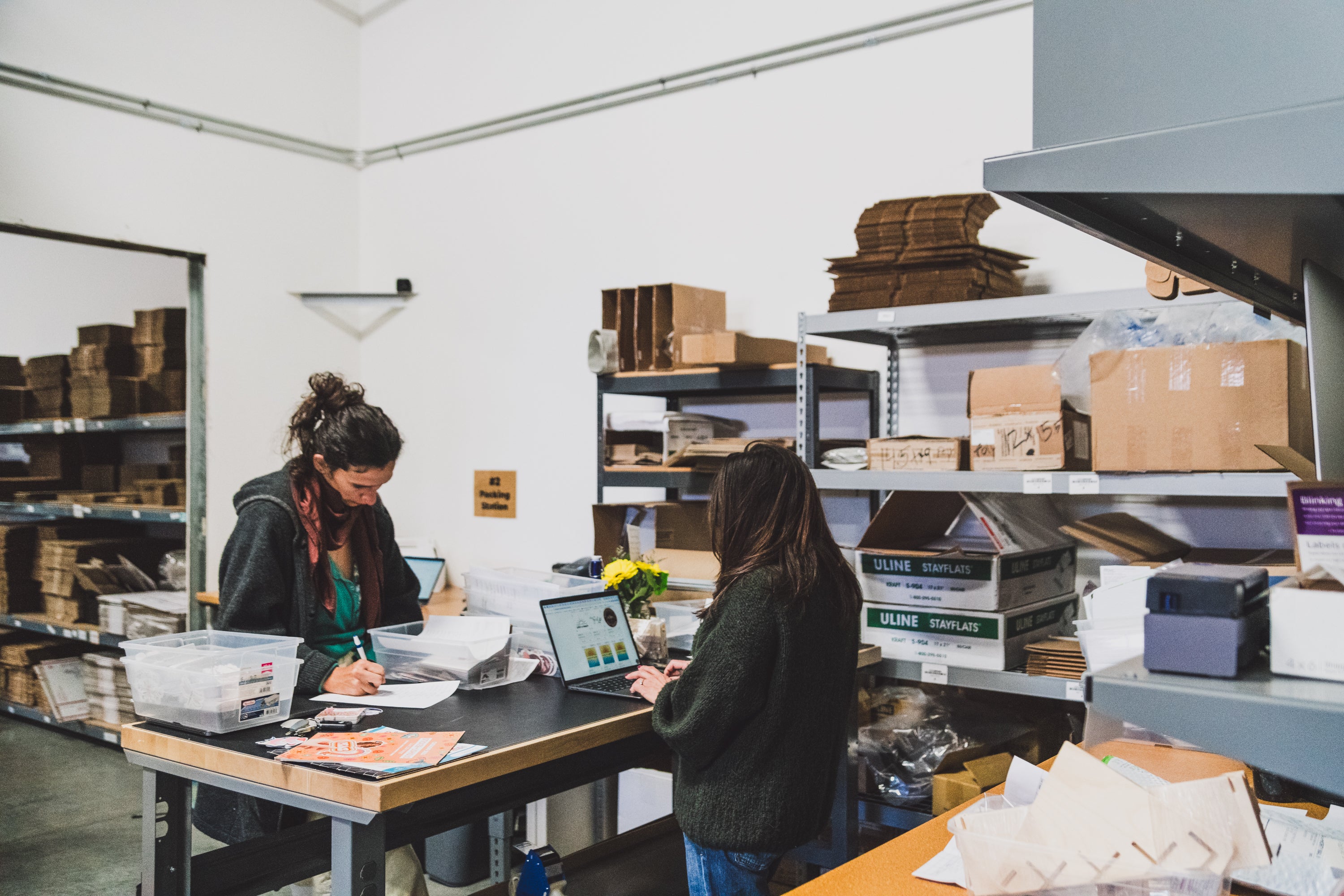 Collective Pallet team members working at a warehouse packing station with inventory shelves, shipping supplies, and order fulfillment materials in the background.