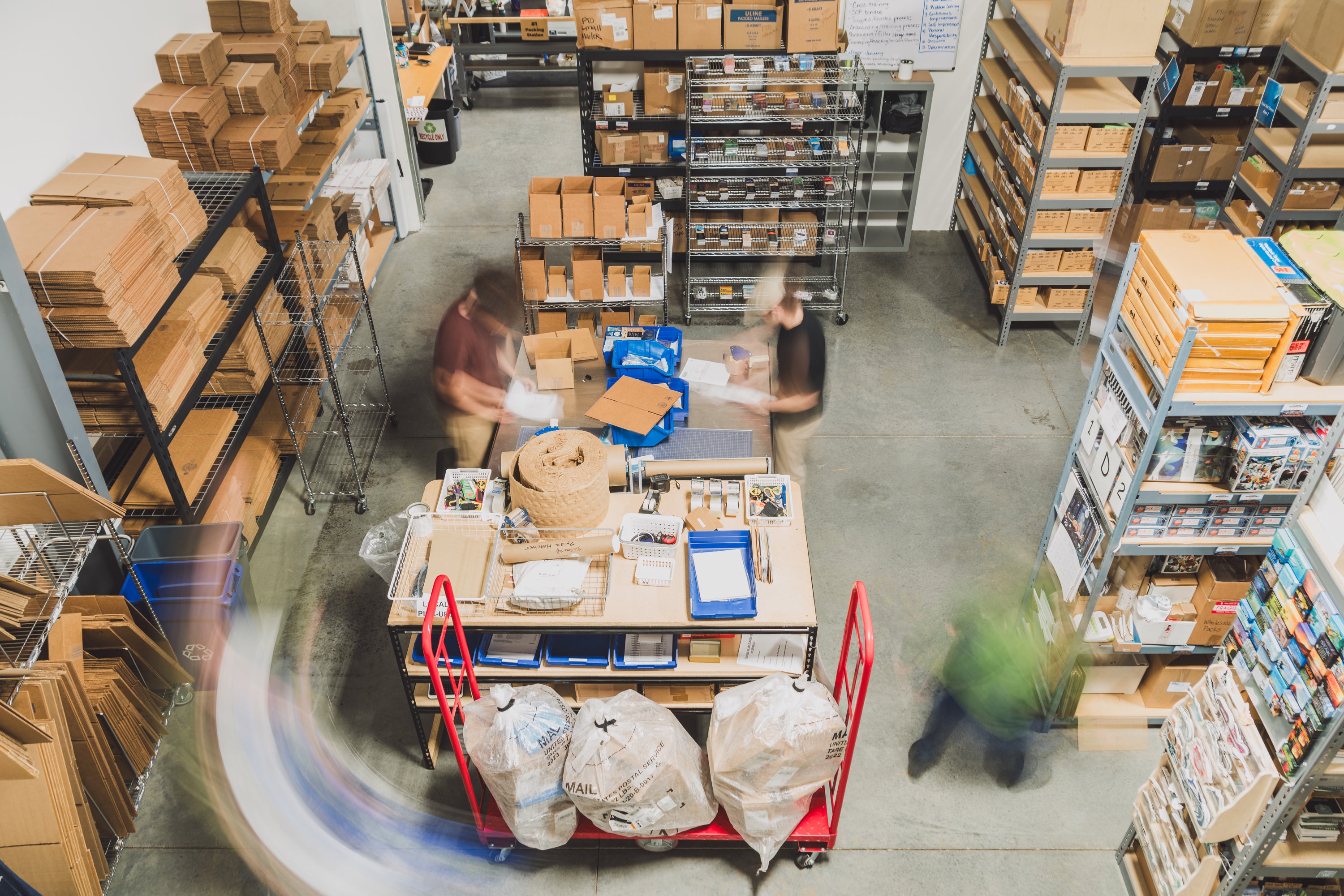 Overhead view of the Collective Pallet warehouse fulfillment workspace with team members packing orders, organized inventory shelving, cardboard shipping materials, and active packing stations.